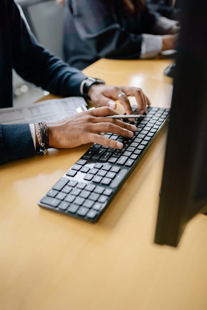 person-holding-a-pencil-while-typing-on-the-black-keyboard-8867210 Close-up of hands typing on keyboard in an office setting, showcasing work professionalism.